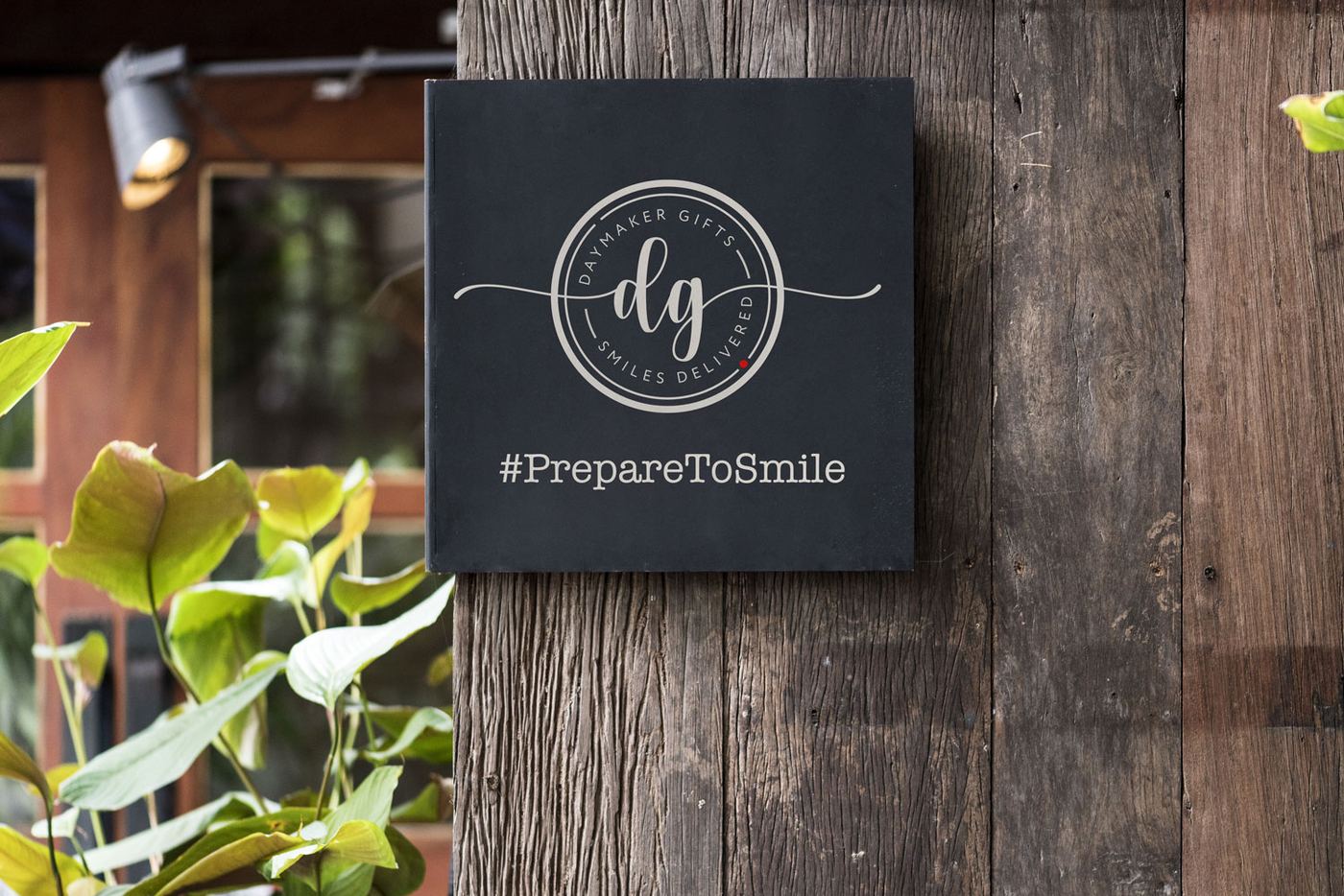 Daymaker Gifts seal on a dark slate sign against reclaimed wood, with the hashtag PrepareToSmile